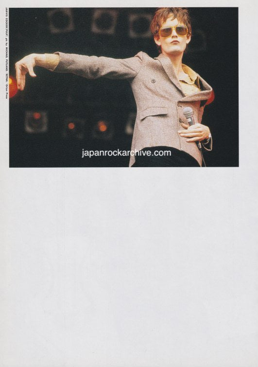 Jarvis Cocker of Pulp in a suit with sunglasses on stage making a unique hand shape