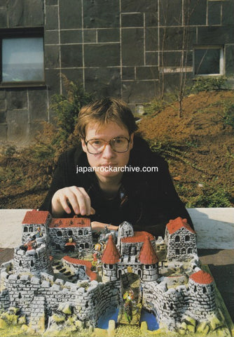 Andy Partridge of XTC with glasses in front of a model of a castle-like structure outdoors.